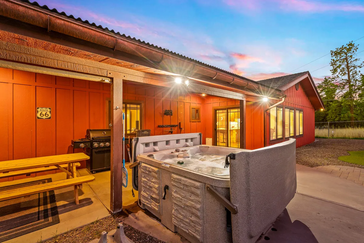 A spacious deck area is shown with an outdoor dining table, a barbecue grill, and a hot tub overlooking the natural landscape. Friends are seen relaxing in the hot tub, barbecuing, and sharing meals outdoors as the sun sets.