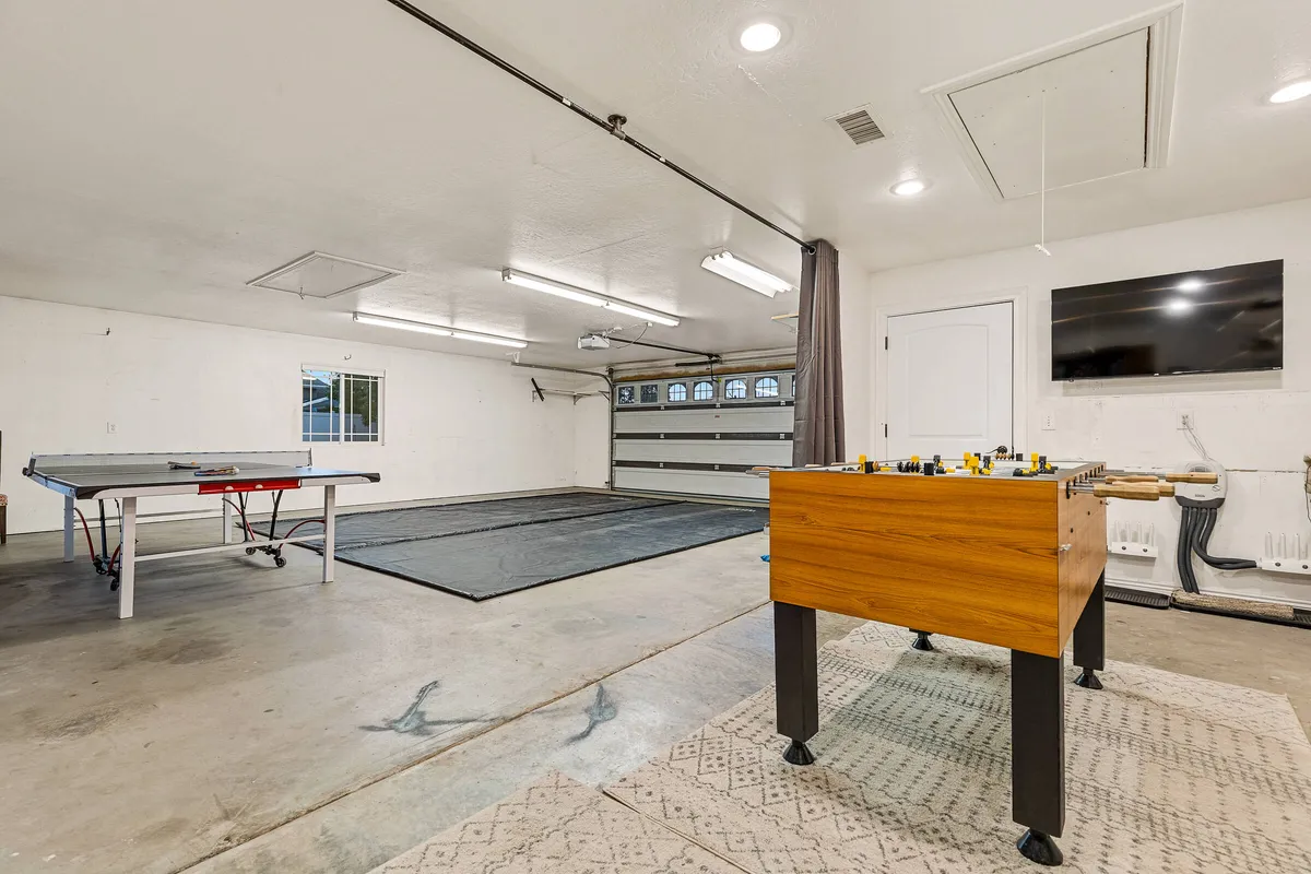 This image showcases a dedicated game room featuring both a ping pong and a foosball table, a garage door that hints at extra space and light, and a TV mounted on the wall, suggesting options for tournaments and group gatherings. The overall setting is lively, casual, and perfect for multi-generational fun.