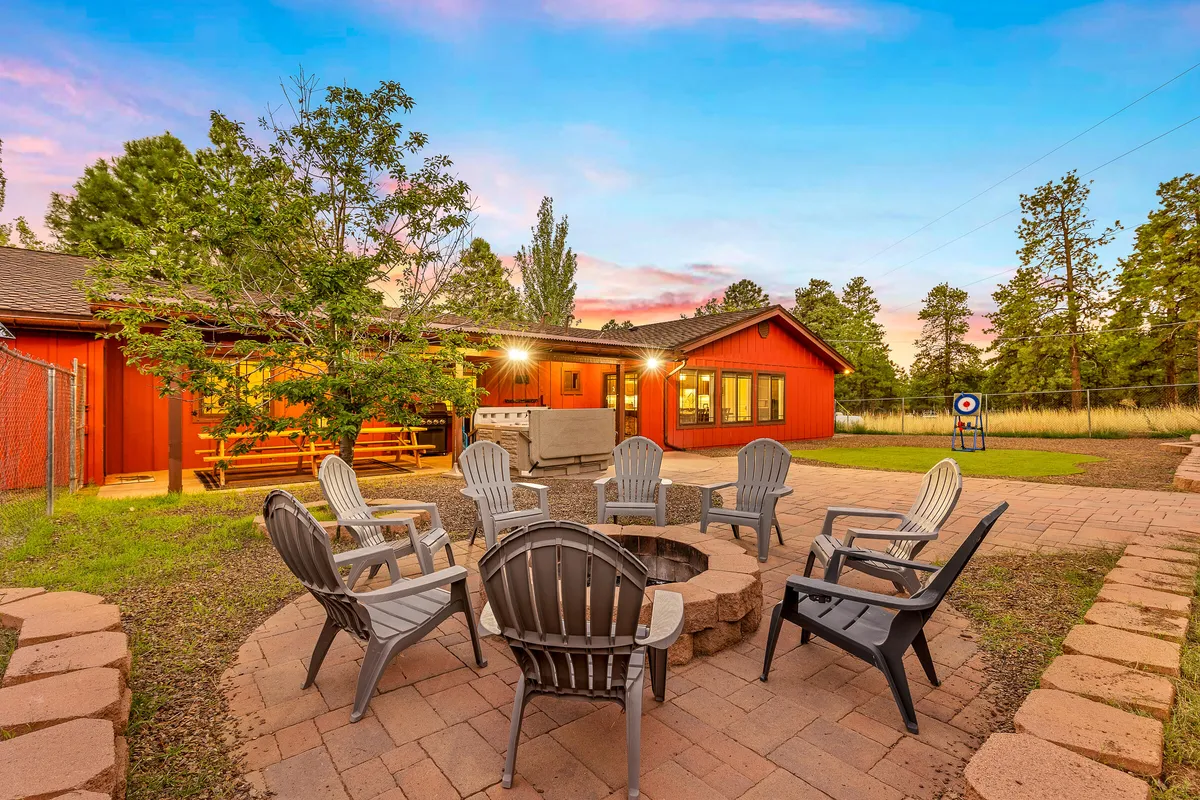 The image depicts a cozy fire pit area with a circle of Adirondack chairs set against the house and framed by surrounding pine trees. People are gathered around the fire, barbecuing, stargazing, and celebrating outdoors, evoking a sense of relaxation and camaraderie.