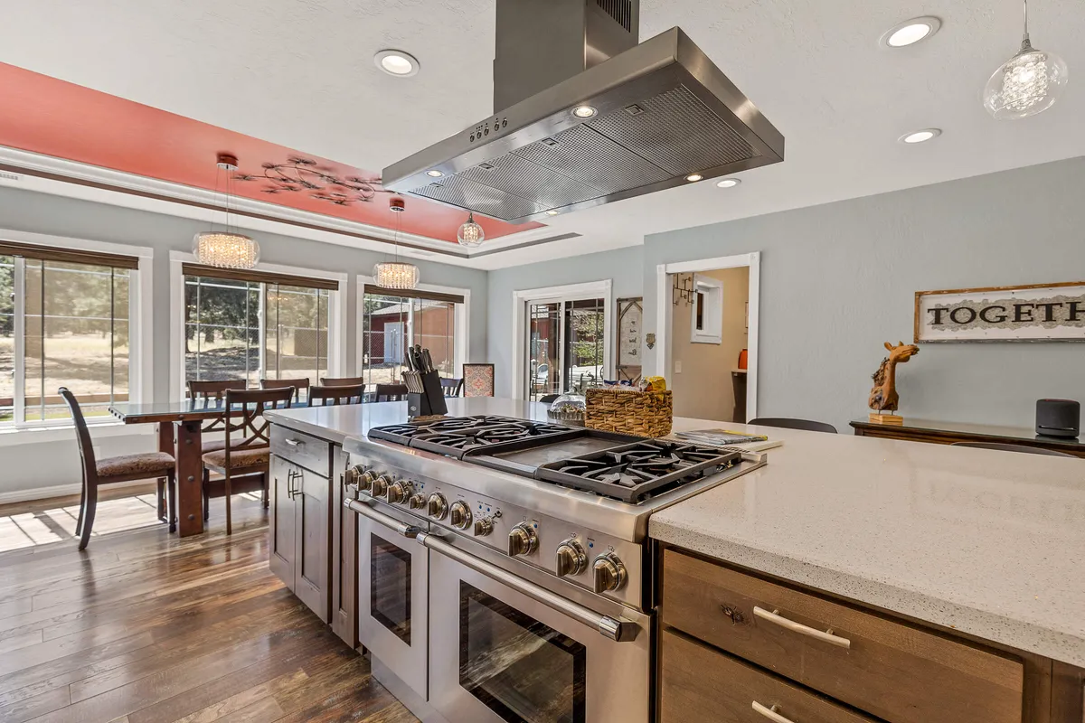 A modern kitchen and dining area bathed in natural light, featuring a large kitchen island, gas range, and dining area beside expansive windows overlooking forest views. The space is ideal for cooking, gathering, and enjoying the scenery.