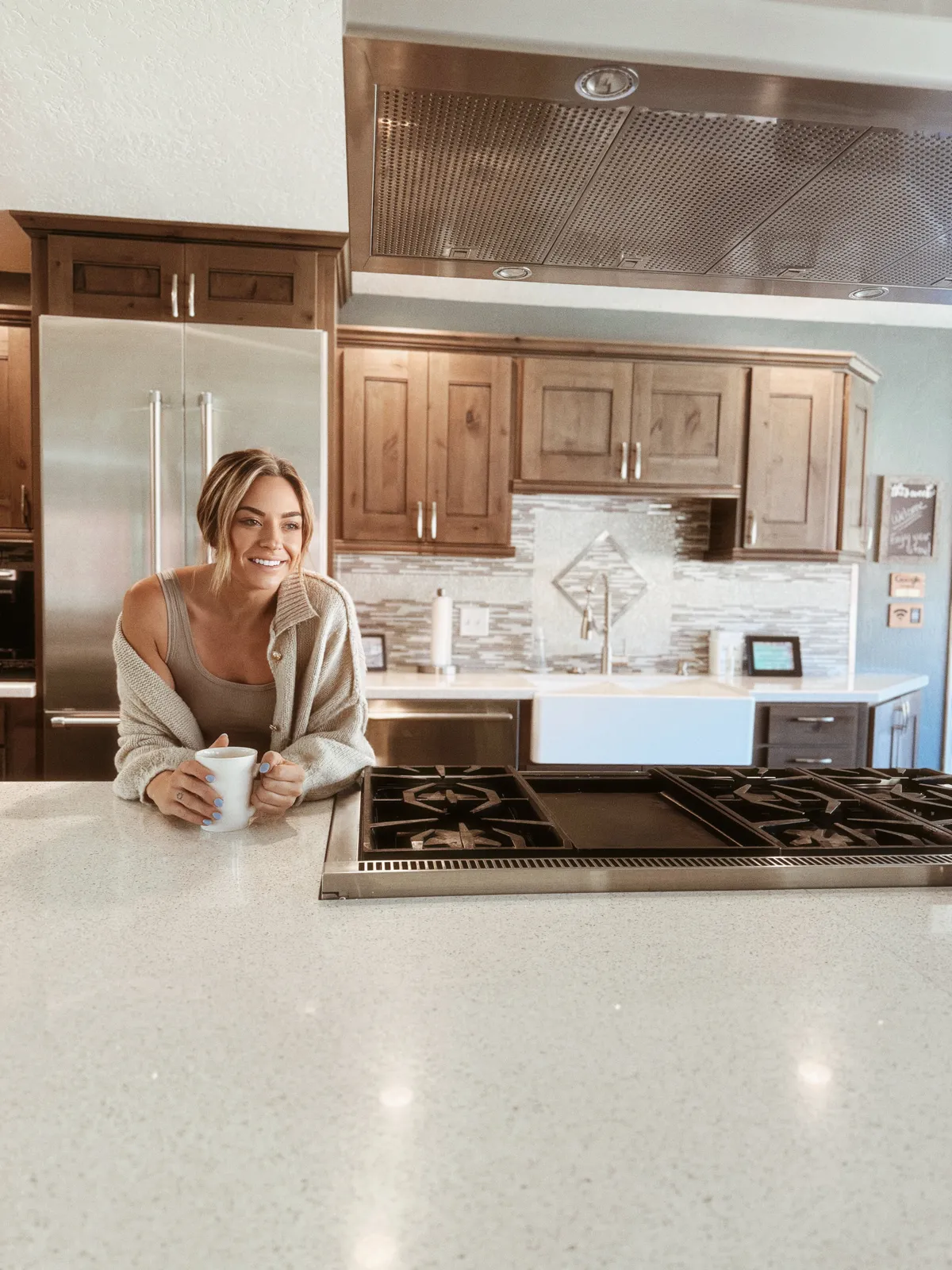 A kitchen scene featuring a prominent kitchen island with a gas cooktop, where a woman enjoys a coffee mug. The atmosphere is relaxed, and the kitchen is well-equipped, suggesting both functionality and comfort for casual breakfasts or festive gatherings.