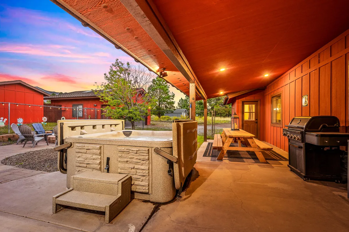 This image features an inviting outdoor setting with a bubbling hot tub surrounded by towering pines. The area also includes a picnic table, barbecue grill, and comfortable outdoor seating. The scene captures people relaxing in the hot tub, preparing food, and socializing, with a beautiful sunset view in the background.