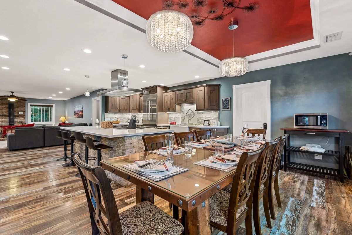 A warm, inviting dining room with a large wooden table set beneath elegant chandeliers. The kitchen island and bar stools are visible in the background, with light streaming in through nearby windows. The setup suggests a space perfect for group meals, celebrations, and gathering with friends or family.