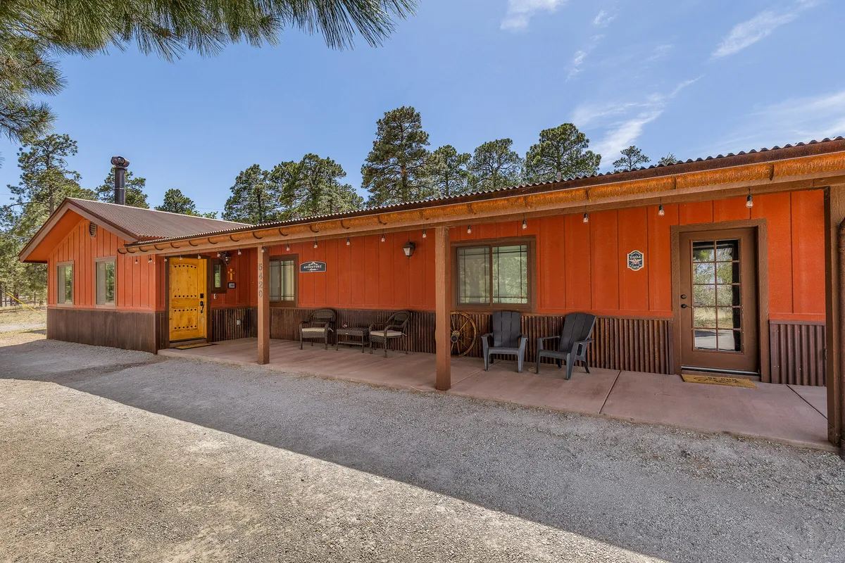 A welcoming rustic orange exterior with a large wooden door, cozy outdoor seating, and mature Ponderosa pine trees surrounding the property. The porch invites relaxation, and the adjacent yard hints at direct access to scenic nature trails.