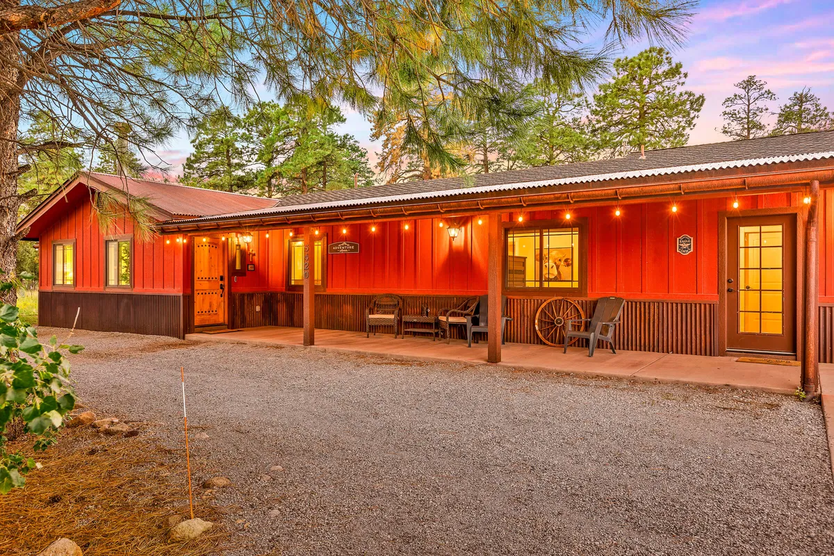 The image showcases the inviting exterior of a single-family home nestled among towering Ponderosa Pines. The wooden front porch is warmly lit by string lights, featuring rustic double doors and a charming wagon wheel decoration. The scene exudes a cozy, welcoming atmosphere perfect for group gatherings. Sunset hues cast a golden glow over the porch, hinting at the promise of starlit evenings and shared outdoor meals.